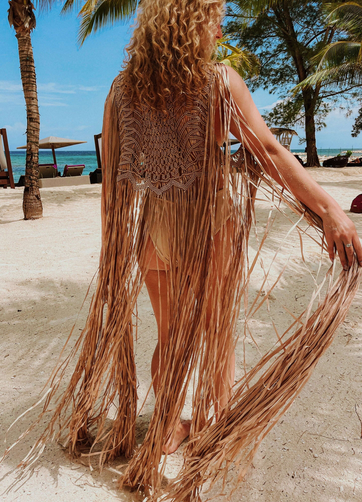 A person with curly hair stands on a sandy beach beneath palm trees, dressed in the Mia Linen Dress and Macrame Cover Up. This flowing ensemble shifts gracefully as they walk, creating a relaxed, tropical mood. In the background are lounge chairs and a view of the sea under a clear blue sky, capturing an elegant beach-to-evening chic style.