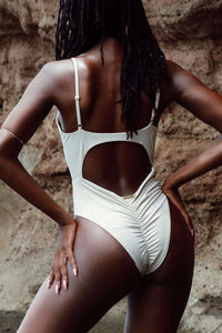 A person with long hair wearing the Mahalia One Piece Swimsuit stands confidently with one hand on their hip, back facing the camera against a rocky background.