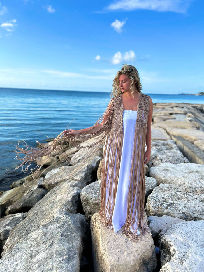 A woman wearing the Mia Linen Dress and Macrame Cover Up stands on rocks by the sea. The sky is a clear blue with a few scattered clouds, and the calm water enhances the perfect beach-to-evening chic vibe.