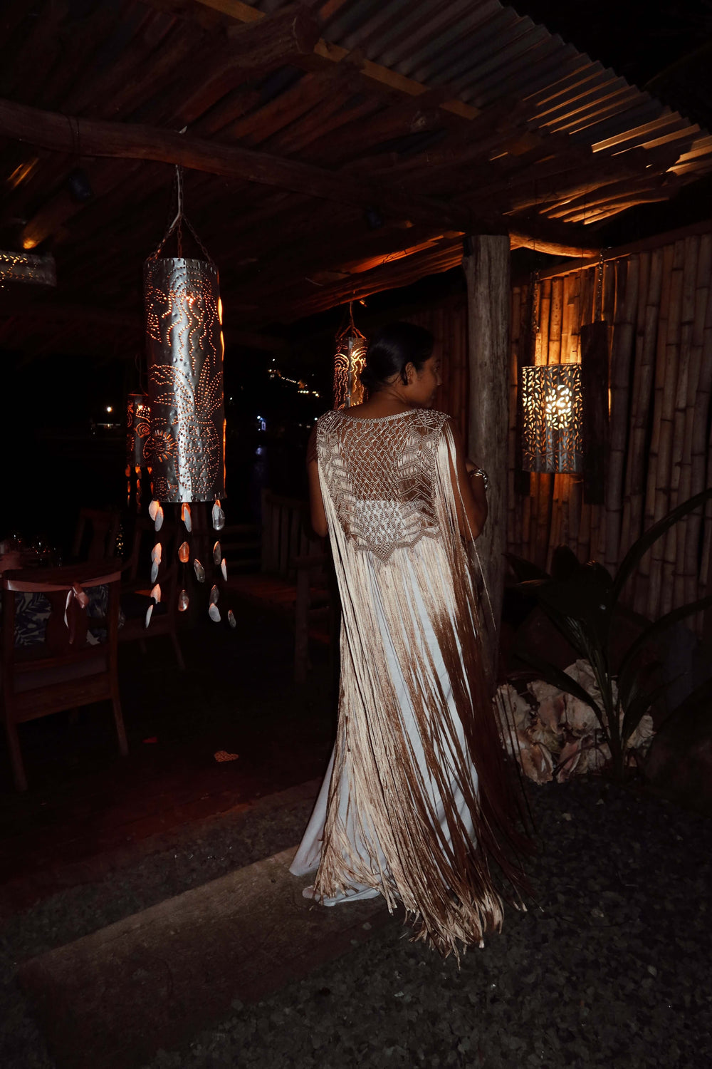 A woman wearing the Mia Linen Dress with a handmade macrame cover-up stands with her back to the camera in a dimly lit, rustic setting. She is surrounded by decorative hanging lights and wooden furniture, creating a warm and cozy ambiance.