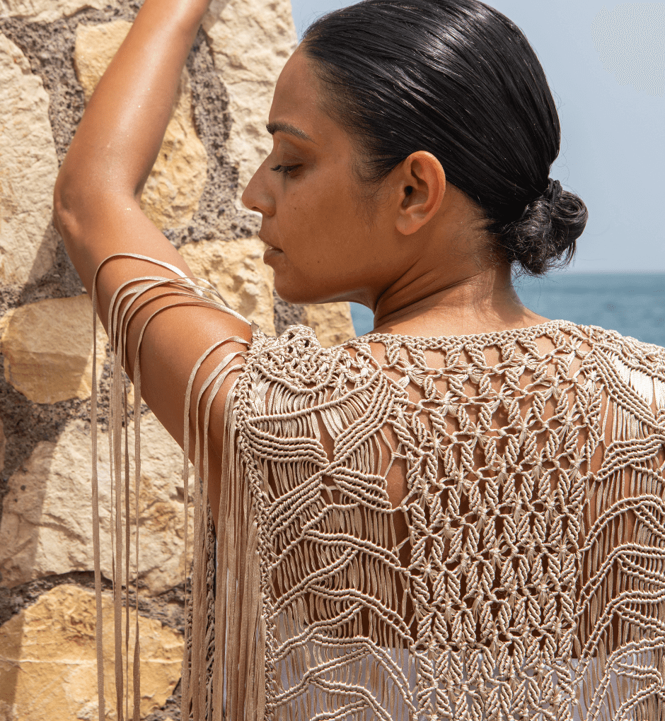 A person with dark hair styled in a bun leans against a stone wall by the sea, wearing the Mia Linen Dress paired with a handmade macramé cover-up featuring intricate patterns and tassels, achieving a bohemian, beach-to-evening chic look.