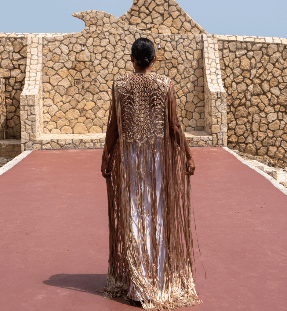 A person stands on a red walkway, their back to the camera, wearing the Mia Linen Dress and Macrame Cover Up in brown and gold. The scene exudes beach-to-evening chic against the backdrop of a stone wall with distinct patterns, set beneath a clear blue sky.