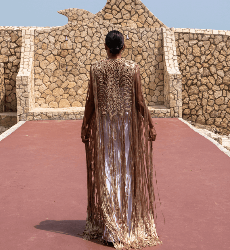 A person stands on a red walkway, their back to the camera, wearing the Mia Linen Dress and Macrame Cover Up in brown and gold. The scene exudes beach-to-evening chic against the backdrop of a stone wall with distinct patterns, set beneath a clear blue sky.