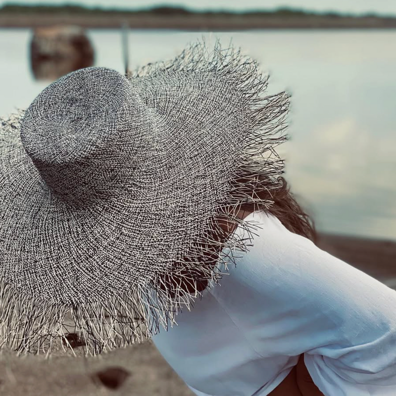 Person wearing a large straw hat on a beach with water in the background