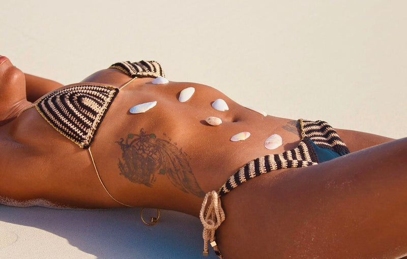 A person is lying on the sand wearing the Celeste Crochet Bikini Top, with shells artfully arranged on their stomach. The focus is on the shells and a tattoo across their midsection, while the sand and sunlight create warm, relaxed vibes reminiscent of the Amalfi Coast.