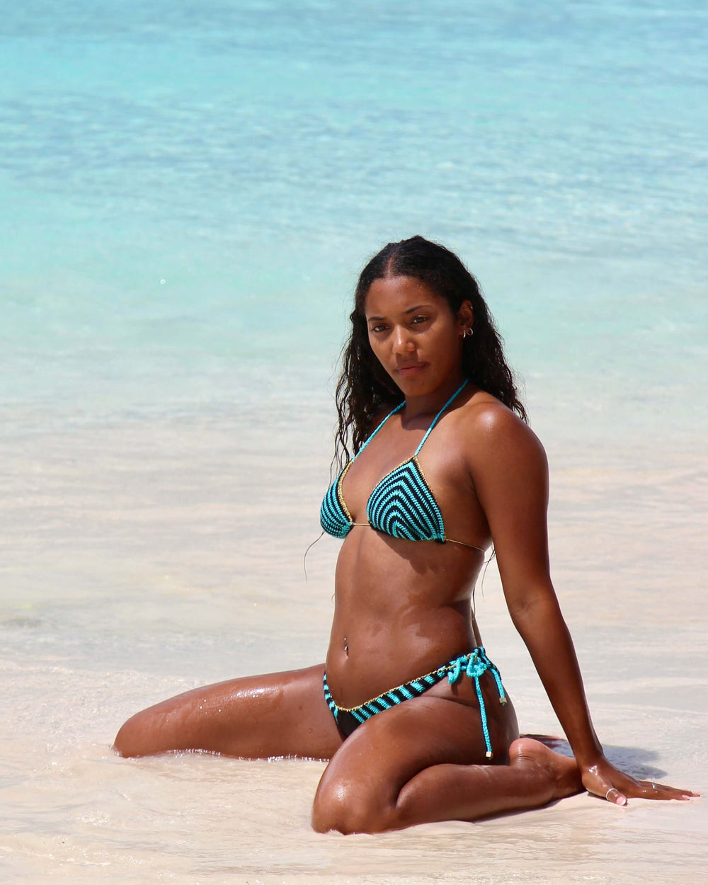 A woman kneels on a sandy beach along the Amalfi Coast, facing the camera. She is wearing the Celeste Crochet Bikini Top. The clear turquoise water gently comes up to her knees, with a bright, sunny sky overhead.