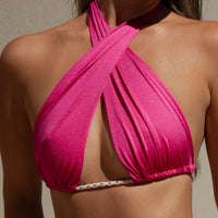 A person is wearing the Hya Convertible Bikini Top, which has a twisted front design in pink and features a halter style. Made from shiny Italian fabric, this bikini top includes adjustable ties and a thin rope-like detail under the bust. They are standing against a light-colored background.